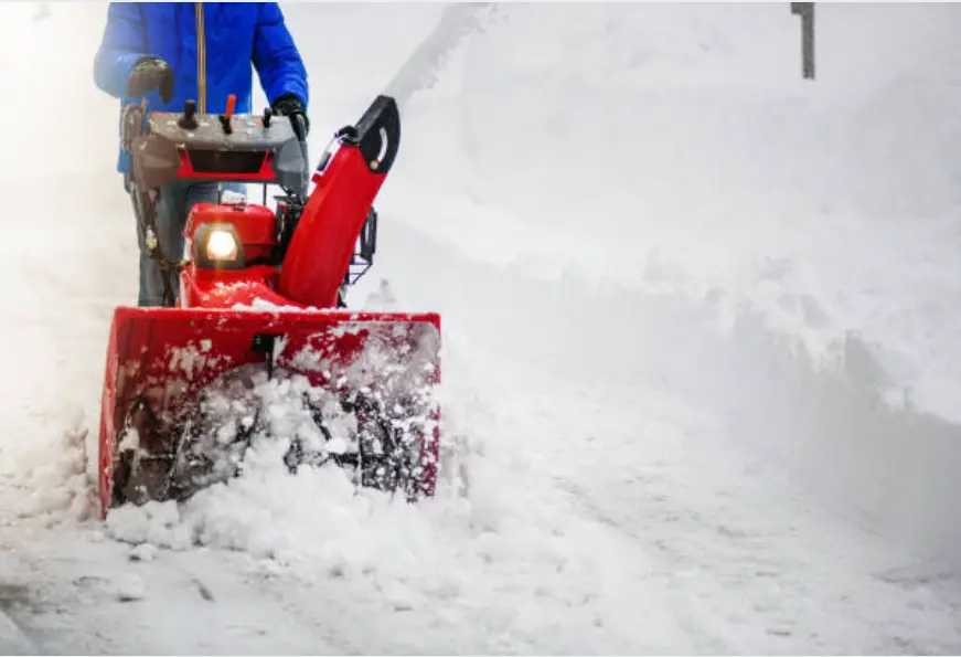 A worker in winter clothing uses a snowblower to clear a snowy walkway.